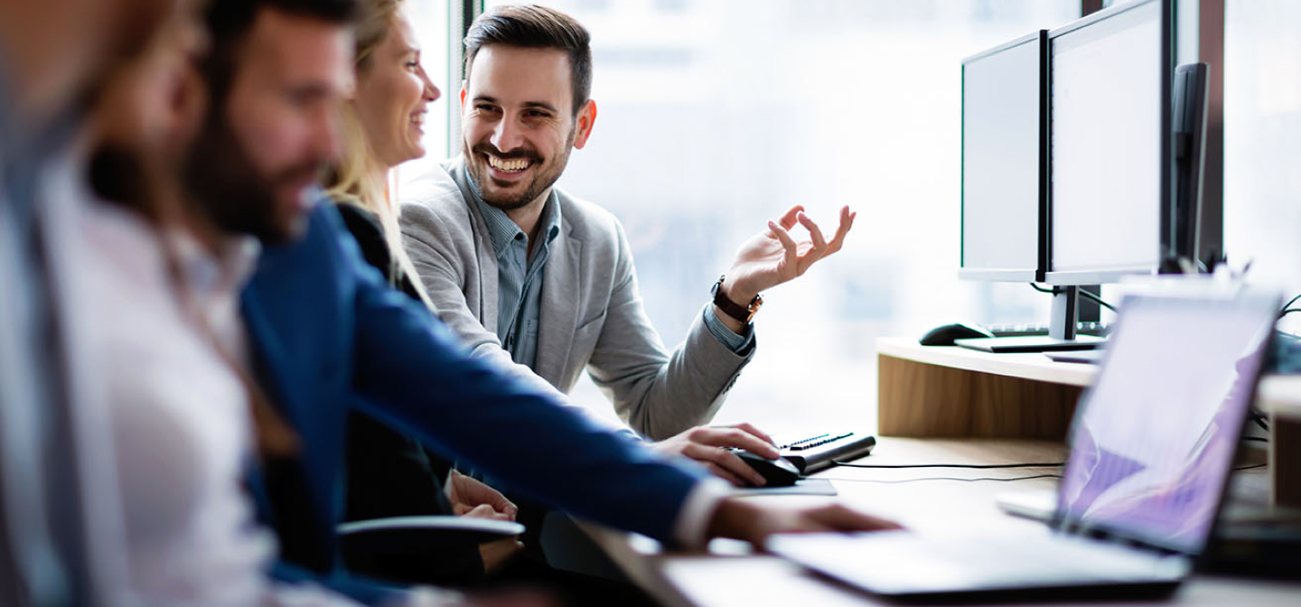 Due persone in secondo piano sorridono e discutono davanti ad un pc con due monitor